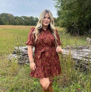 Woman in a red patterned dress and brown boots standing in a field with trees in the background